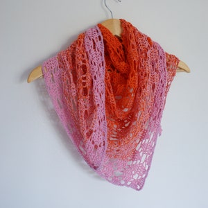 May include: A crocheted shawl with a pink and orange ombre color gradient. The shawl is hanging on a wooden hanger against a white wall.