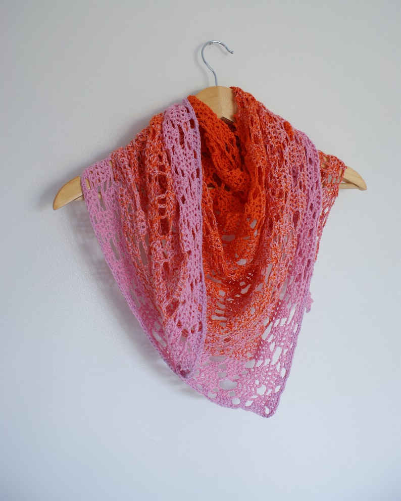 May include: A crocheted shawl with a pink and orange ombre color gradient. The shawl is hanging on a wooden hanger against a white wall.