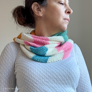 May include: A colorful crochet cowl in a geometric pattern. The cowl features blocks of white, pink, teal, green, and yellow. The handmade accessory is worn around the neck and is made of soft yarn.