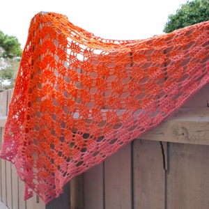 May include: A crocheted shawl with a gradient of orange and pink colors. The shawl is made with a delicate openwork pattern and is draped over a wooden fence.