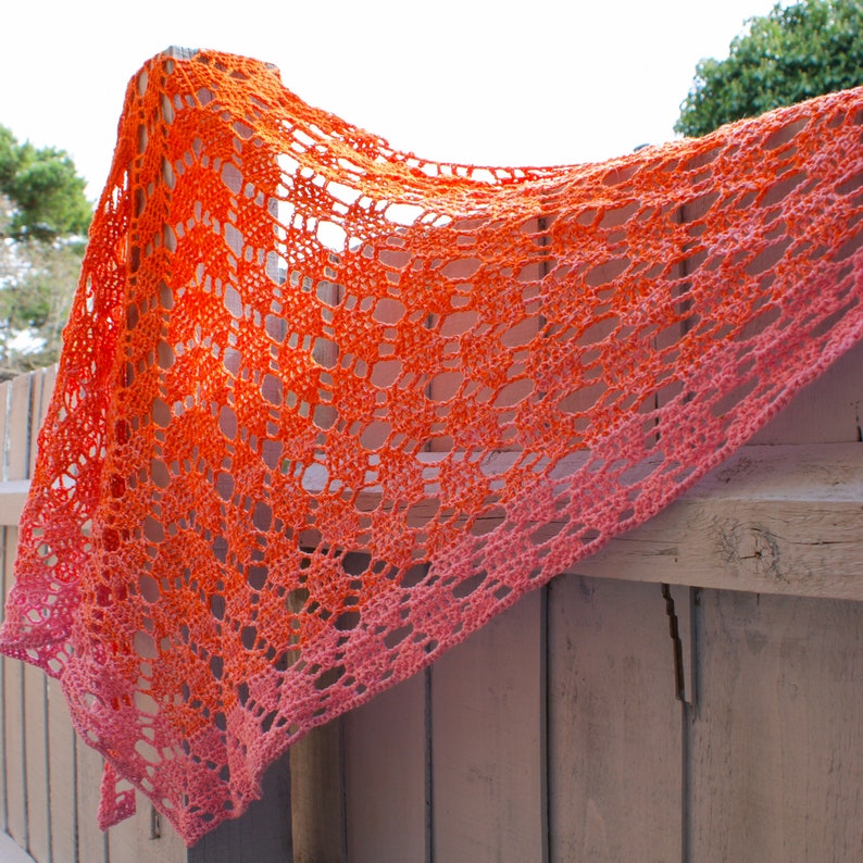 May include: A crocheted shawl with a gradient of orange and pink colors. The shawl is made with a delicate openwork pattern and is draped over a wooden fence.