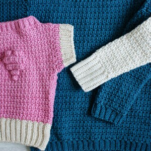 May include: Three crocheted sweaters in different colors and patterns. One is pink with a white trim and a heart-shaped design, another is teal with a white trim, and the third is white with a teal trim.
