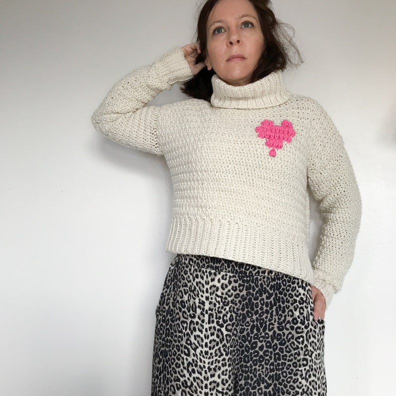 May include: A white crocheted turtleneck sweater with a pink crocheted heart design. The sweater is cropped and has long sleeves. The model is wearing the sweater with a leopard print skirt.