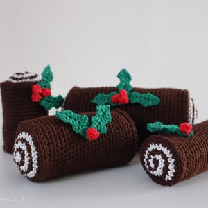 May include: Four crocheted yule log ornaments in various sizes, decorated with green holly leaves and red berries. The logs are brown with white frosting details.