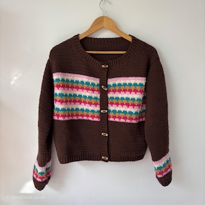 May include: A dark brown crochet cardigan featuring a horizontal band of colourful geometric patterns in pink, teal, and yellow across the chest and sleeves. The garment has a round neckline, long sleeves, and five light-coloured wooden buttons.