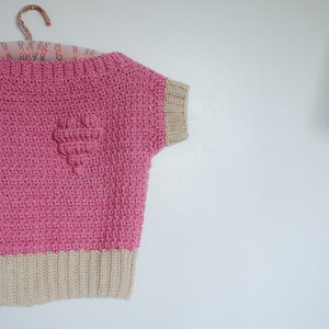 May include: A pink crocheted top with a beige crocheted trim and a heart-shaped design on the front.