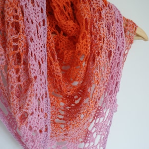 May include: A crocheted shawl with a gradient of pink and orange colors. The shawl is made with a delicate lace pattern and is draped over a wooden hanger.