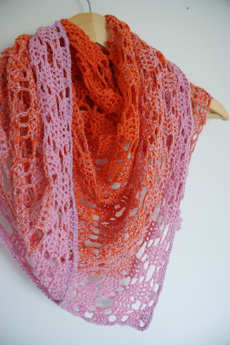 May include: A crocheted shawl with a gradient of pink and orange colors. The shawl is made with a delicate lace pattern and is draped over a wooden hanger.