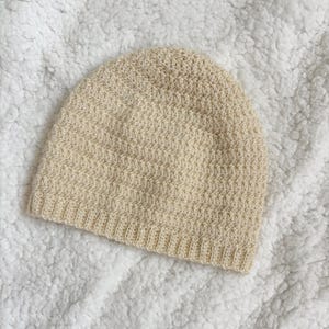 May include: A cream-colored, hand-knitted beanie hat. The hat features a textured stitch pattern and a ribbed brim. The soft, neutral color makes it a versatile accessory for various outfits. The hat is likely made of a warm, comfortable material.