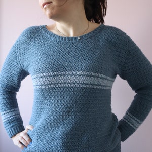 A woman stands hands on hips, proudly wearing a blue long sleeve, round neck textured crochet sweater. The crochet jumper has 3 horizontal stripes around the chest and each wrist on the long sleeves.