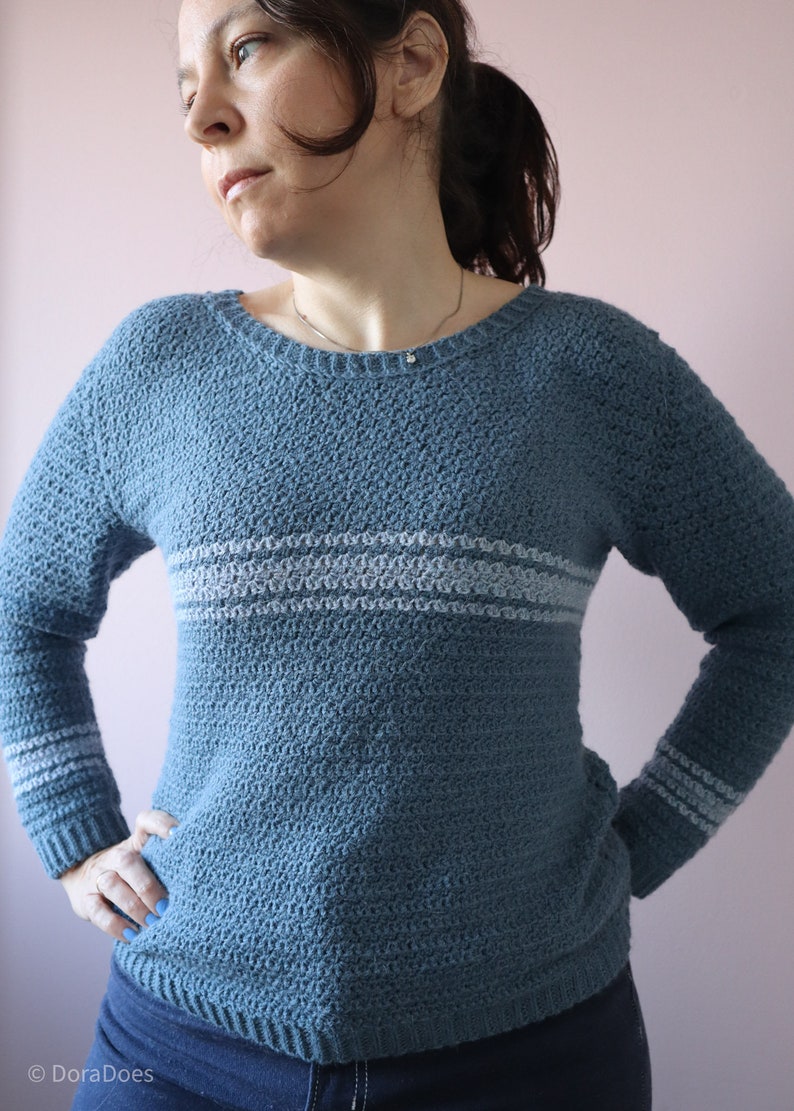 A woman stands hands on hips, proudly wearing a blue long sleeve, round neck textured crochet sweater. The crochet jumper has 3 horizontal stripes around the chest and each wrist on the long sleeves.