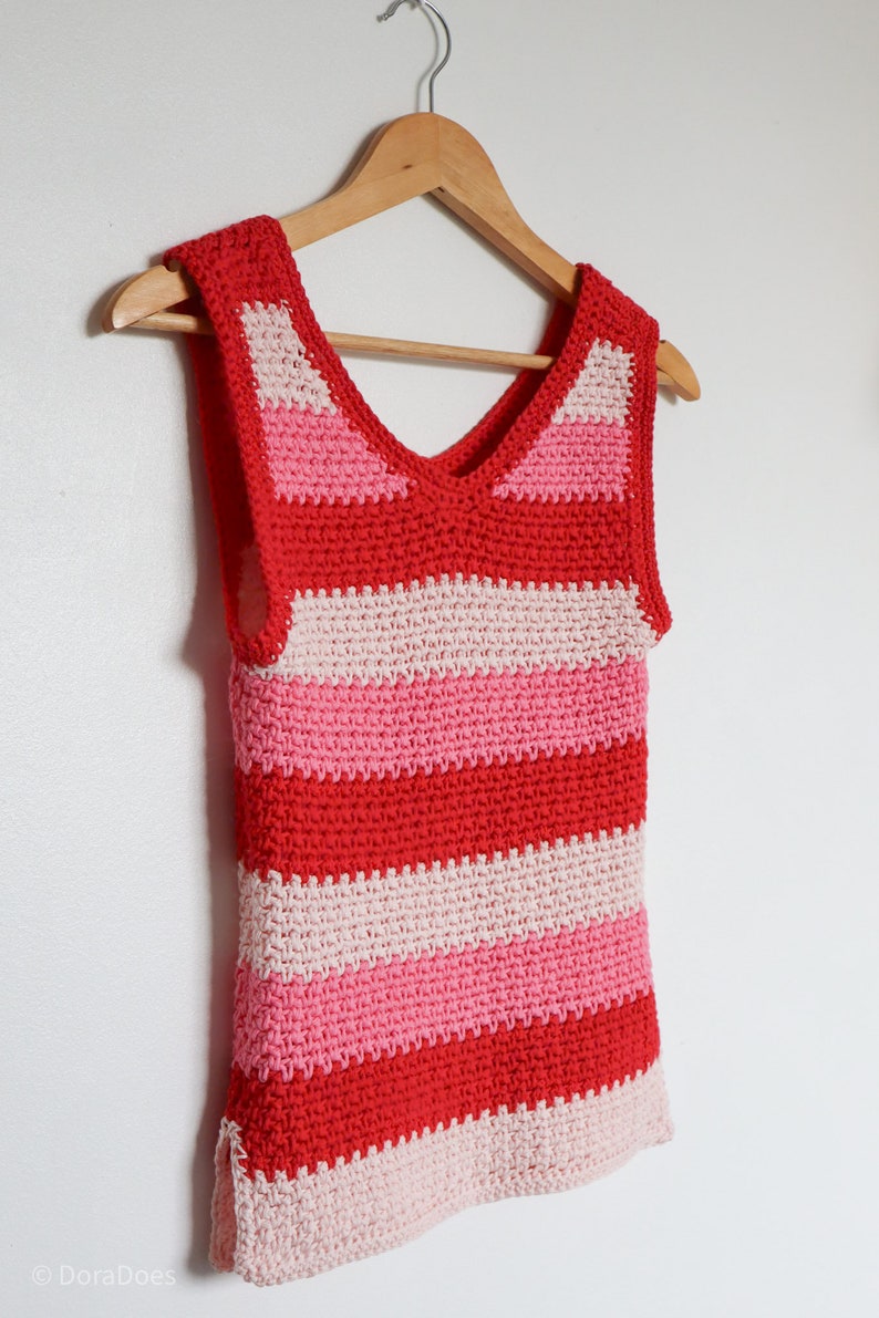 A crochet tank top made in bright cotton aran weight yarn is seen from the side as it hangs on a white wall.