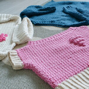 May include: Three crocheted sweaters in different colors: white with a pink heart, pink with a white trim, and blue. The sweaters are laid out on a textured surface.