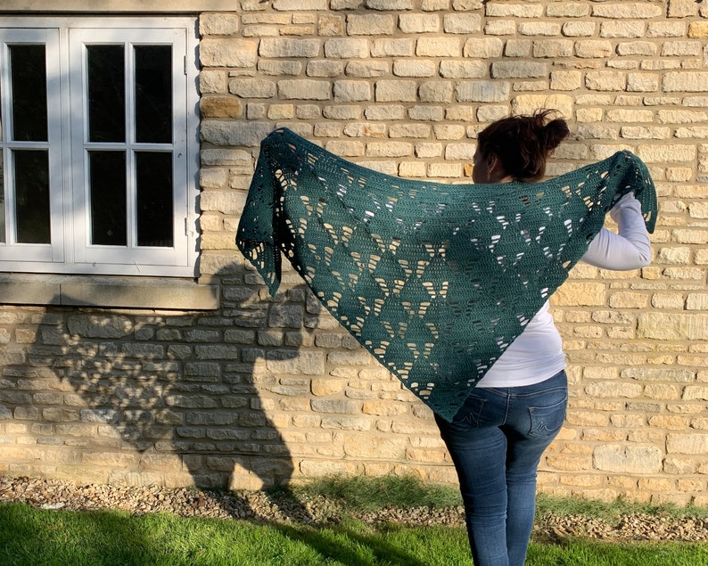 May include: A green crocheted shawl with a lacy pattern. The shawl is draped over the shoulders of a person standing in front of a stone wall.