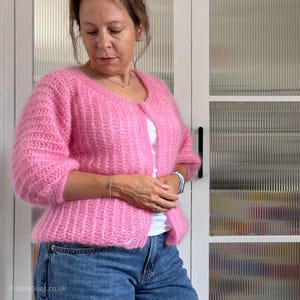 May include: A pink, open-front cardigan with a textured knit design. The cardigan has three-quarter sleeves and a slightly flared hem. The model is wearing the cardigan over a white top and blue jeans. The cardigan is made of a soft, fluffy yarn.
