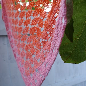 May include: A crocheted shawl with a gradient of orange and pink colors. The shawl is made with a delicate lace pattern and is hanging from a branch.