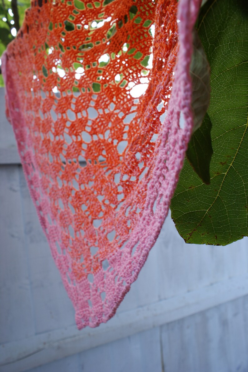 May include: A crocheted shawl with a gradient of orange and pink colors. The shawl is made with a delicate lace pattern and is hanging from a branch.