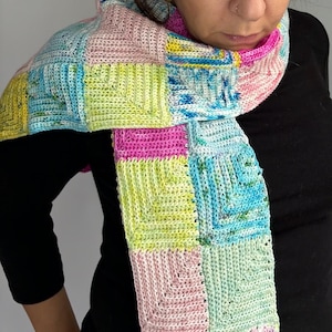 May include: A colorful crocheted scarf with squares in shades of pink, green, blue, and yellow. The scarf is made with a textured stitch and has a fringed edge.