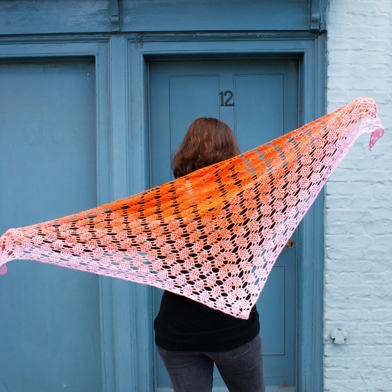 May include: A woman wearing a crocheted shawl with a gradient of orange to pink. The shawl is draped over her shoulders and she is standing in front of a door with the number 12 on it.
