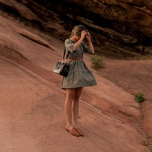 May include: A woman wearing a black and white gingham dress with a brown leather bag and brown loafers stands on a red rock surface.