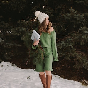Puede incluir: Una mujer con un gorro de punto blanco y un vestido verde con volantes y hombros descubiertos sostiene un regalo envuelto frente a un árbol de hoja perenne nevado.