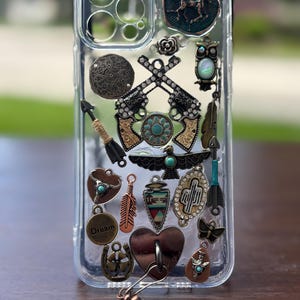 May include: Clear phone case adorned with various charms, including crossed pistols, feathers, a horseshoe, and a heart. The charms feature turquoise and metallic accents, creating a bohemian or Western aesthetic.