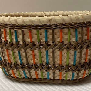 Puede incluir: Una cesta ovalada tejida con una base marrón claro y un patrón de rayas marrones, verdes, naranjas y azules.