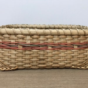 May include: A rectangular woven basket with a natural brown color and a decorative band of red, green, and brown woven into the top edge.