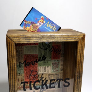 May include: A wooden box with a glass front, decorated with vintage ticket stubs and the word "TICKETS" in black lettering. The box has a slot on the top for inserting items. A ticket for "Le Nouba Cirque du Soleil" is partially visible in the slot.