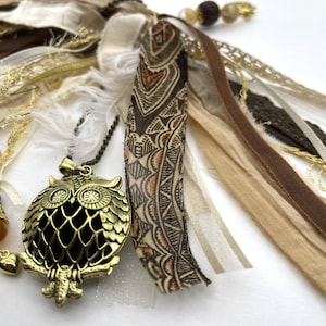 May include: A gold owl charm pendant with a delicate, intricate design hangs from a brown chain. The pendant is surrounded by a variety of brown, beige, and white ribbons and lace.