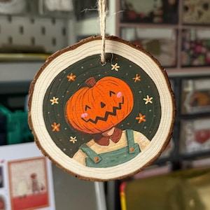 May include: A wooden round ornament with a painted image of a person with a pumpkin for a head. The pumpkin is orange with a carved jack-o'-lantern face. The person is wearing a green shirt and overalls. The background is green with white stars and flowers.
