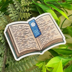 May include: A sticker depicting an open book with a blue bookmark. The bookmark features a moon and stars design and the text "THIS IS WHERE I FELL ASLEEP". The book's pages are light brown, and the sticker has a white border.