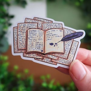 Könnte beinhalten: Ein Aufkleber mit einem Stapel gealterter Papierbögen mit einem offenen Buch oben. Das Buch hat ein Pentagramm und handschriftlichen Text. Eine dunkelviolette Federkiel liegt auf den Seiten, mit Tintenklecksen. Der Aufkleber hat einen weißen Rand.