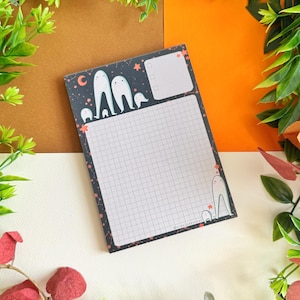 May include: A notepad with a dark blue cover featuring a ghost design, stars, and a crescent moon. The notepad has a grid pattern and a section for checkboxes. The notepad is surrounded by green and red foliage.
