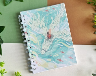 Może przedstawiać: Spiralny notatnik z fantastyczną ilustracją białego smoka i jeźdźca na turkusowo-żółtym tle. Okładka zawiera czerwone ptaki i płynny wzór. Notatnik otoczony jest zielonymi liśćmi.