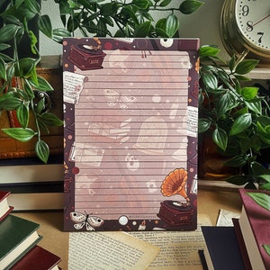 Pode incluir: Um bloco de notas com linhas com uma estética vintage que apresenta um toca-discos, uma borboleta e um gramofone. O bloco de notas tem um fundo castanho com um padrão de riscas rosa claro e branco.