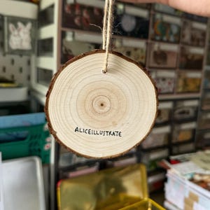 May include: A round wooden ornament with a natural wood grain finish. The ornament has a hole at the top for hanging and the text "Aliceillustrate" is written on the front.