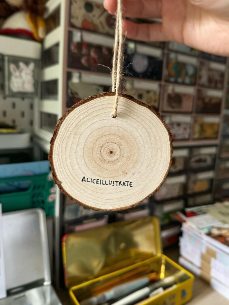 May include: A round wooden ornament with a natural wood grain finish. The ornament has a hole at the top for hanging and the text "Aliceillustrate" is written on the front.