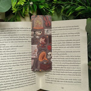 May include: A bookmark with a vintage aesthetic featuring a variety of illustrations, including a butterfly, a skull, a gramophone, a violin, and a glass dome with a plant inside. The bookmark is printed on a dark background with a light brown border.