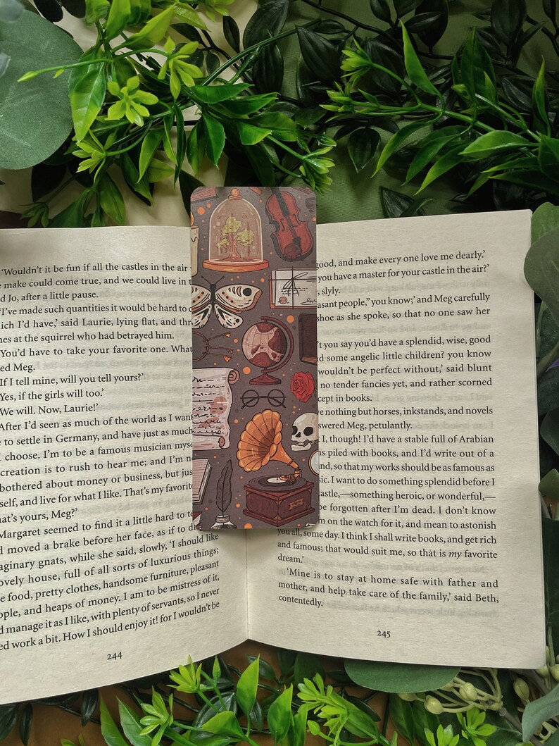 May include: A bookmark with a vintage aesthetic featuring a variety of illustrations, including a butterfly, a skull, a gramophone, a violin, and a glass dome with a plant inside. The bookmark is printed on a dark background with a light brown border.