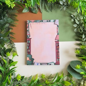 May include: A notepad with a pink background and a library-themed border. The border features bookshelves, a green armchair, and scattered books. The text "notes from the library..." is at the top. The notepad is surrounded by green foliage.