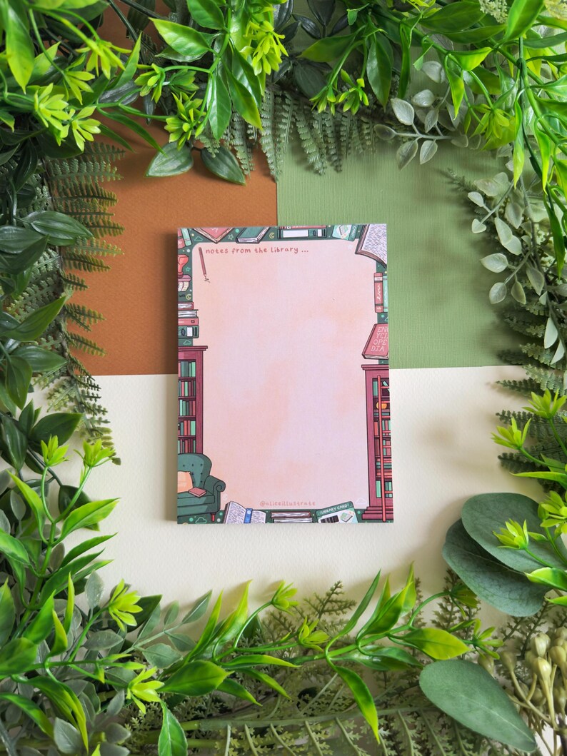 May include: A notepad with a pink background and a library-themed border. The border features bookshelves, a green armchair, and scattered books. The text "notes from the library..." is at the top. The notepad is surrounded by green foliage.