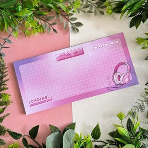 Pode incluir: Um bloco de notas rosa e roxo com um padrão de grade e um gráfico de barra de carregamento. O bloco de notas tem o texto "LEVEL UP" e um gráfico de fones de ouvido.