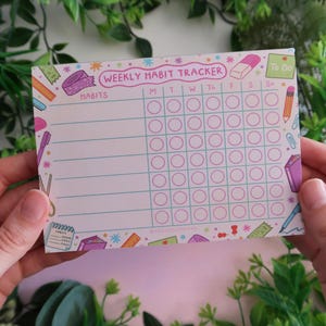 Puede incluir: Un bloc de notas semanal para el seguimiento de hábitos con un encabezado rosa que dice WEEKLY HABIT TRACKER. La cuadrícula incluye columnas para los días de la semana y filas para los hábitos, decorada con ilustraciones coloridas de artículos de papelería.