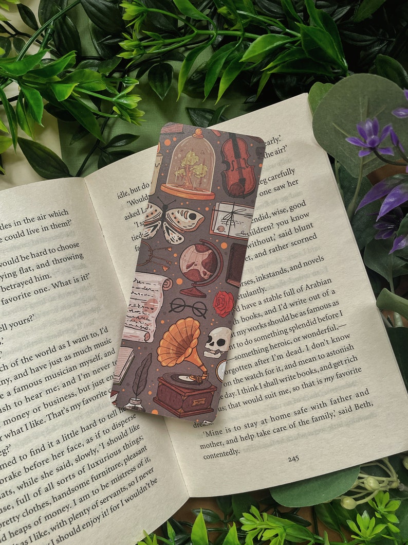 May include: A bookmark featuring a vintage aesthetic with illustrations of a butterfly, a globe, a gramophone, a skull, a violin, a rose, a book, and a glass dome with a tree inside. The bookmark is on a book with text that reads "Mine is to stay at home safe with father and mother, and help take care of the family." said Beth, contentedly.