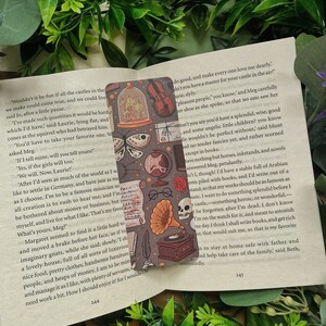 May include: A book bookmark with a dark gray background and a repeating pattern of illustrations. The illustrations include a butterfly, a skull, a gramophone, a violin, and a glass dome with a bird inside. The bookmark is placed on a page of a book with text about castles and dreams.