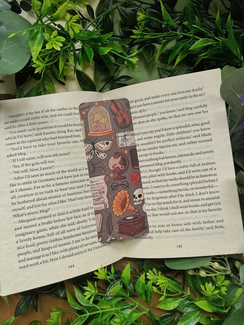 May include: A book bookmark with a dark gray background and a repeating pattern of illustrations. The illustrations include a butterfly, a skull, a gramophone, a violin, and a glass dome with a bird inside. The bookmark is placed on a page of a book with text about castles and dreams.