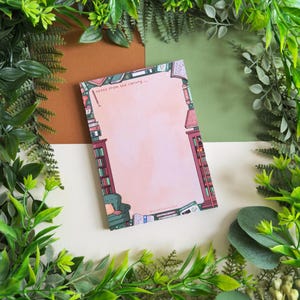 May include: A rectangular notepad with a pink center and a library-themed border. The border features illustrations of bookshelves, a green armchair, and scattered books. The notepad has the text "notes from the library..." at the top.