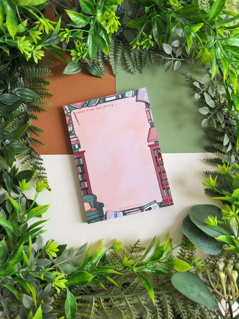 May include: A rectangular notepad with a pink center and a library-themed border. The border features illustrations of bookshelves, a green armchair, and scattered books. The notepad has the text "notes from the library..." at the top.