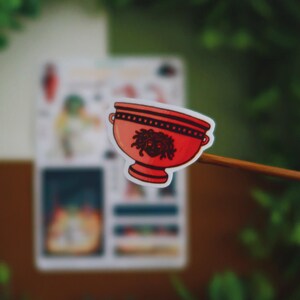 May include: A red sticker featuring a stylized bowl with a Medusa head design. The bowl has a black rim and handles. The sticker is held against a blurred background of green foliage and a white paper.